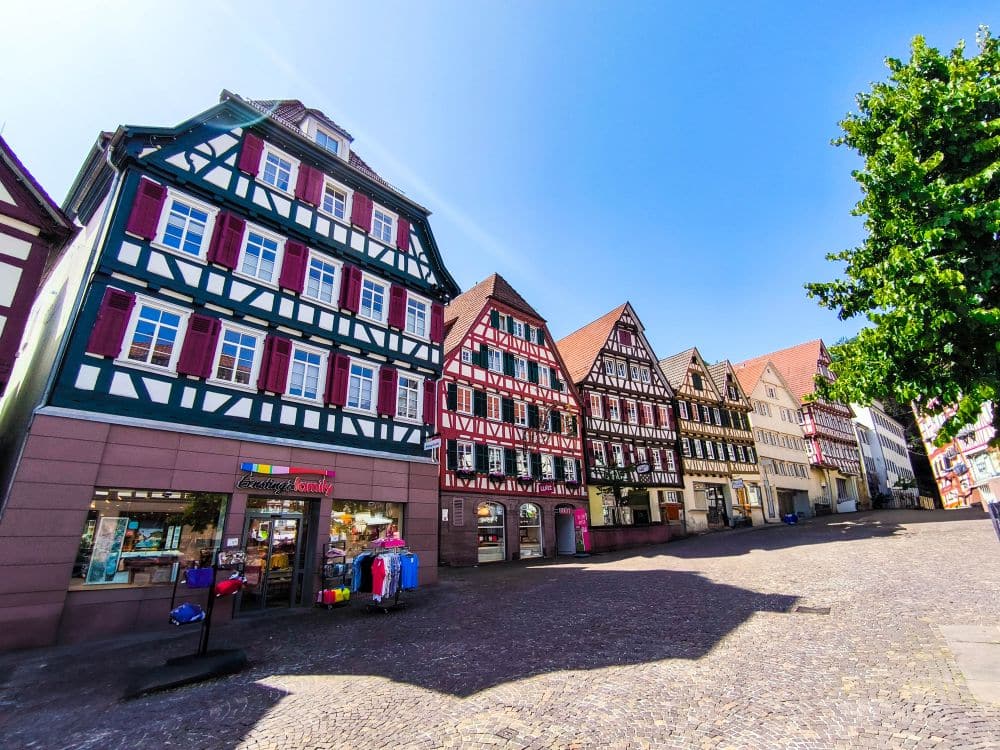 Bunte Fachwerkhäuser säumen eine Kopfsteinpflasterstraße in Calw bei strahlend blauem Himmel, mit einem Geschäft, in dem Kleidung ausgestellt wird, und einem belaubten Baum am rechten Rand des Bildes.