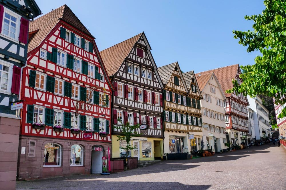 Eine Reihe farbenfroher Fachwerkhäuser mit steilen Giebeldächern säumen eine Kopfsteinpflasterstraße in Calw unter einem klaren blauen Himmel, wobei auf der rechten Seite des Bildes grüne, belaubte Äste zu sehen sind.
