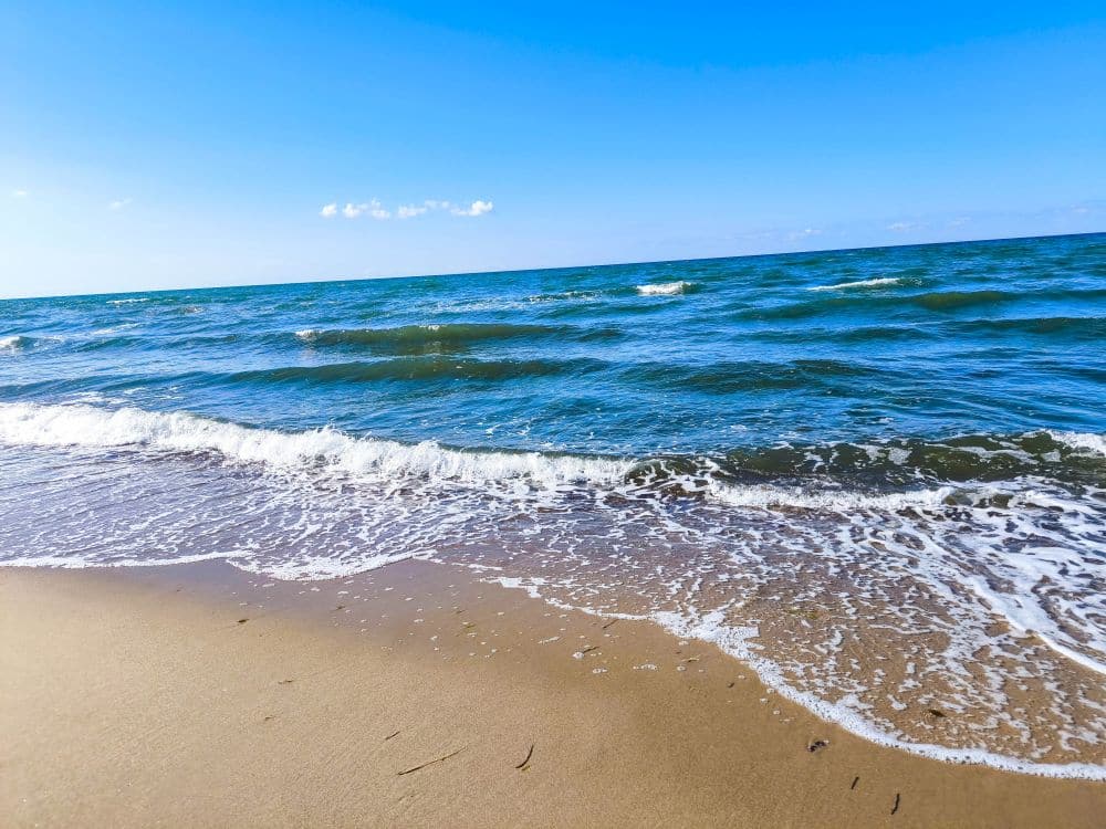 Sanfte Wellen rollen an den Sandstrand von Kühlungsborn unter einem klaren blauen Himmel mit ein paar Wolken am Horizont. Die türkisfarbene Ostsee wirkt ruhig, im Vordergrund glitzert der nasse Sand - perfekt für eine Auszeit am Meer.