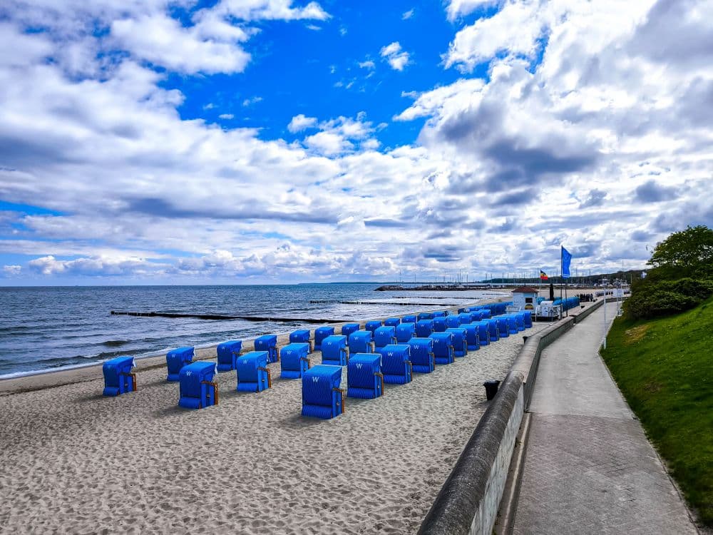 Ein Sandstrand in Kühlungsborn mit blauen Liegestuhlreihen an der Ostsee, rechts ein gepflasterter Weg, darüber ein wolkenverhangener Himmel. Die leere Uferlinie ist sanft geschwungen und lädt zu einer ruhigen Auszeit am Meer ein.