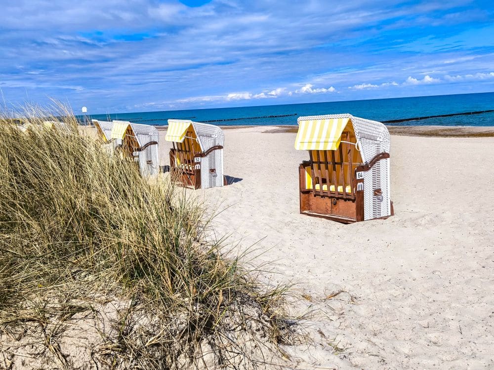 Mehrere weiße Strandkörbe mit gelb gestreiften Schirmen stehen am sandigen Ostseestrand neben hohen Gräsern und bieten eine perfekte Auszeit mit Blick auf Kühlungsborns blaues Meer und den teilweise bewölkten Himmel.