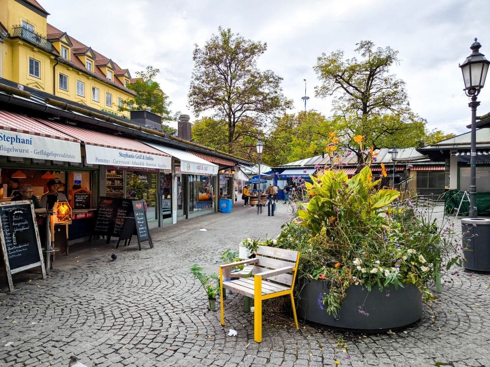 Marktszene im Freien in München mit Essensständen, Kopfsteinpflaster und einer Bank neben einem großen Blumenkübel, der überquillt. Die Einkäufer erkunden die Stadt unter einem wolkenverhangenen Himmel, gelbe Gebäude bilden die charmante Münchner Kulisse.
