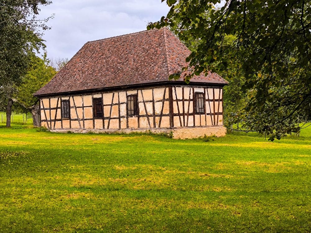 Ein kleines, altes Fachwerkhaus mit Ziegeldach steht auf einer grünen Wiese, umgeben von Bäumen, die an die Gärten und die barocke Pracht in Ludwigsburg erinnern, und das alles unter einem bewölkten Himmel.