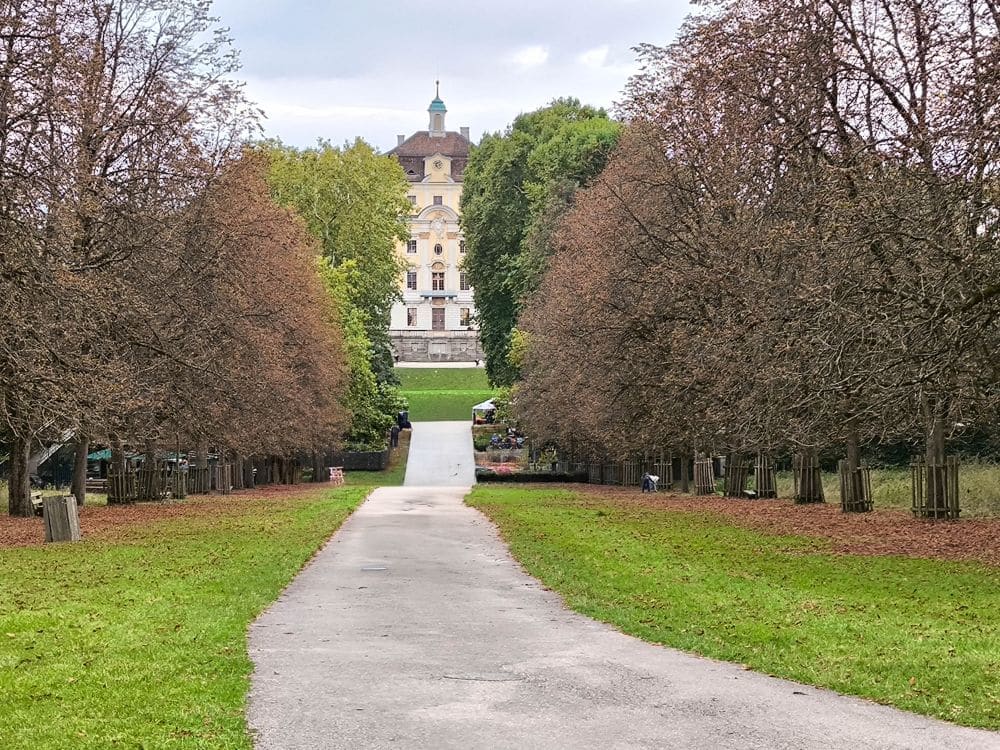 Ein gepflasterter Weg führt durch Reihen von braun belaubten Bäumen in Richtung des großen Ludwigsburger Prunkbaus mit begrüntem Dach, umgeben von Barocke Pracht und üppigem Grün unter einem wolkenverhangenen Himmel.