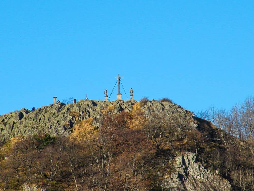 Eine felsige Kuppe in der Rhön, mit drei Statuen und einem großen Kreuz auf dem Gipfel der Milseburg, unter strahlend blauem Himmel, umgeben von blattlosen Bäumen und trockenen Grasflächen - perfekt für ein Wanderabenteuer.