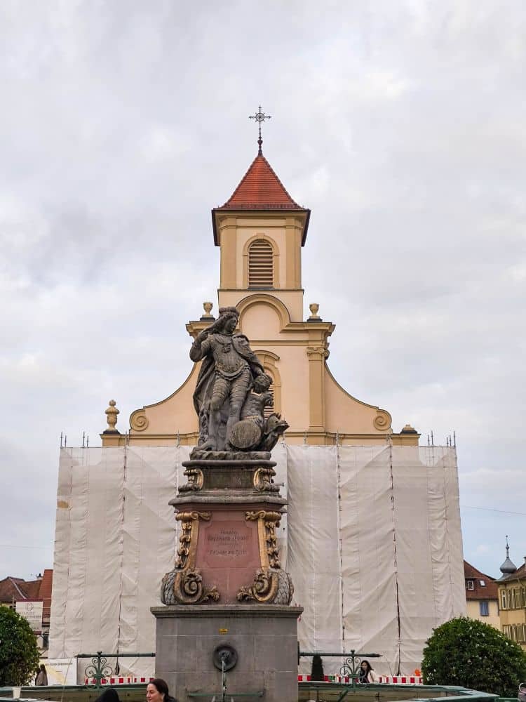 Eine barocke Statue steht vor einer Kirche mit einem rot gedeckten Glockenturm in Ludwigsburg. Die Kirchenfassade ist teilweise von einem weißen Gerüst verdeckt, während der Himmel bedeckt ist und im Hintergrund einige Gebäude zu sehen sind.