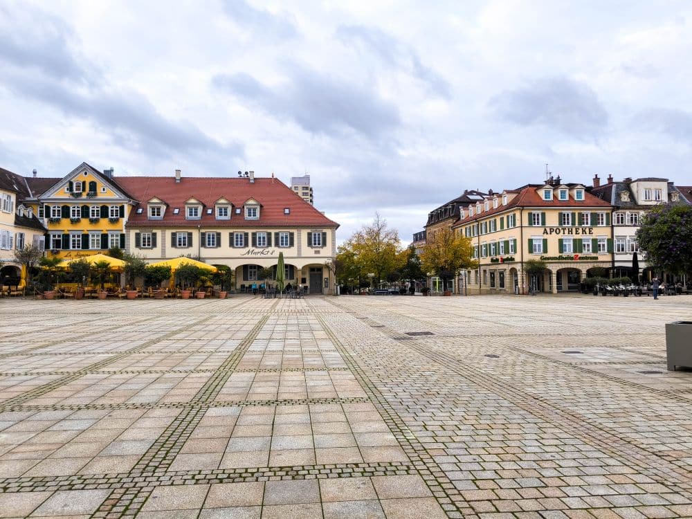 Ein großer, leerer Stadtplatz mit gemusterten Steinfliesen in Ludwigsburg, gesäumt von bunten, traditionellen europäischen Gebäuden mit roten Dächern und Geschäften, unter einem bewölkten Himmel.