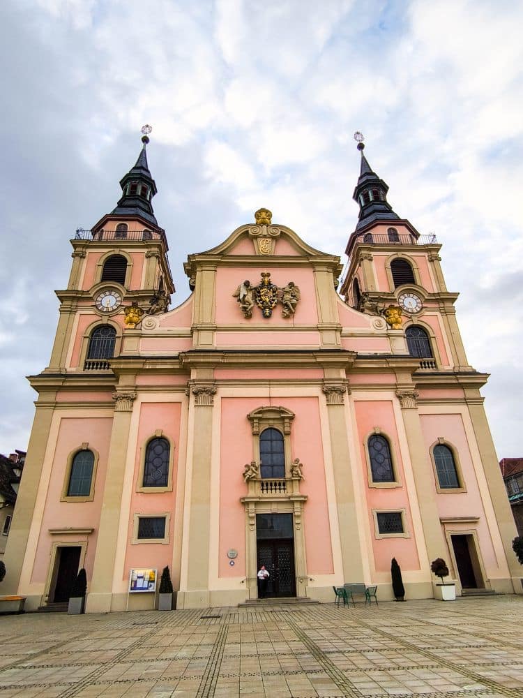 Eine prächtige Barockkirche mit zwei Glockentürmen, rosafarbener und cremefarbener Fassade, Bogenfenstern und kunstvollen Verzierungen steht auf dem weitläufigen gepflasterten Platz von Ludwigsburg unter einem teilweise bewölkten Himmel.