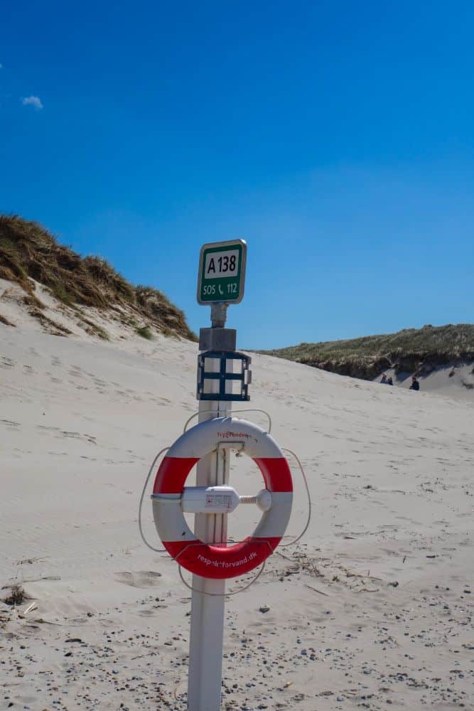 Eine rot-weiße Rettungsboje mit einem grünen SOS-Schild mit der Aufschrift A 138 ist an einem Pfahl am wildromantischen Sandstrand von Blavand in Dänemarks Westen befestigt, vor dem Hintergrund grasbewachsener Dünen und eines klaren blauen Himmels.