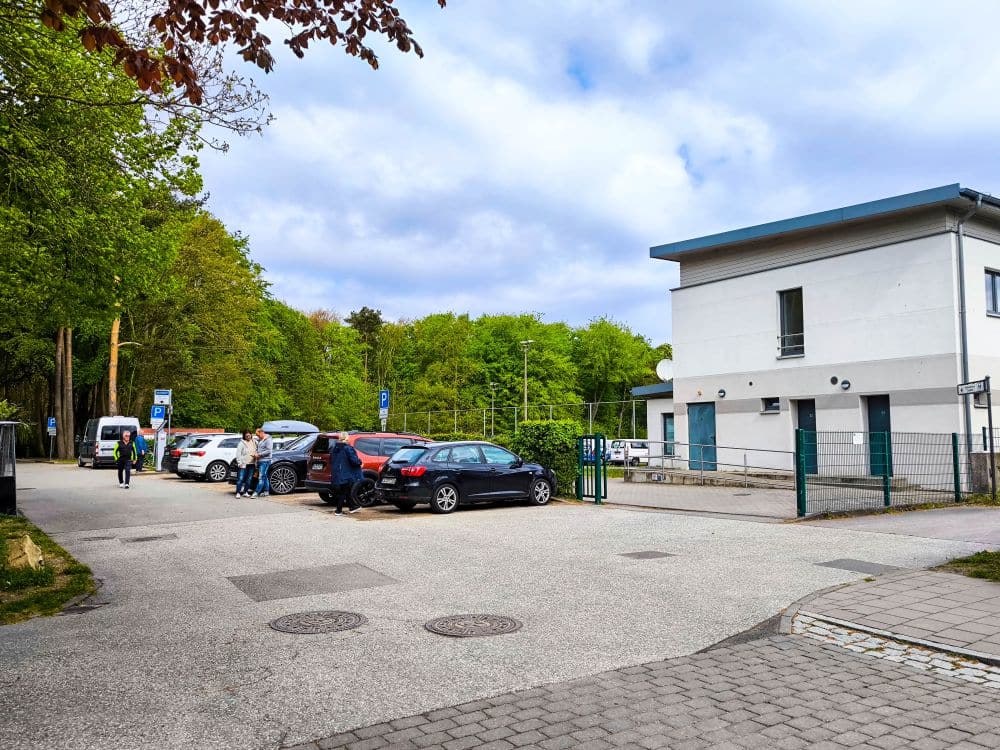 Ein kleiner Parkplatz mit mehreren geparkten Autos, Menschen in der Nähe von Fahrzeugen und ein weißes zweistöckiges Gebäude auf der rechten Seite in Kühlungsborn, umgeben von grünen Bäumen unter einem teilweise bewölkten Ostseehimmel - ein perfekter Ort für eine Auszeit.