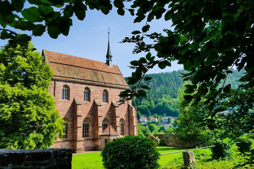 Eine historische Steinkirche mit hohen Bogenfenstern und einem kleinen Kirchturm steht in Hirsau inmitten von üppigem Grün. Im Hintergrund sind bewaldete Hügel und ein paar Häuser unter einem strahlend blauen Himmel zu sehen.