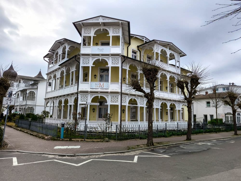 Ein großes, verziertes, dreistöckiges gelb-weißes Gebäude in Binz, mit umlaufenden Balkonen, dekorativen Geländern und blattlosen, beschnittenen Bäumen, die den Bürgersteig unter einem bewölkten Himmel säumen.