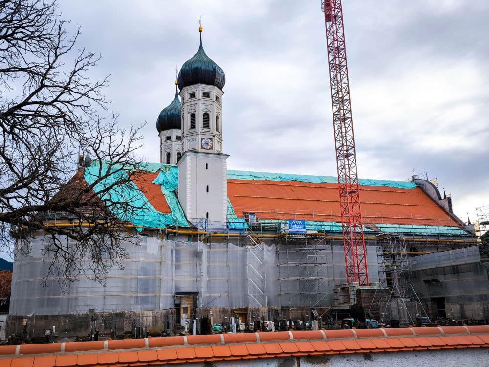 Eine große Barockkirche, das Kloster Benediktbeuern, mit zwei Zwiebeltürmen wird gerade renoviert, bedeckt mit Gerüsten und grünen Planen. Ein roter Baukran steht daneben, vor einem bedeckten Himmel und fernen Bergen. Auf der linken Seite steht ein blattloser Baum.