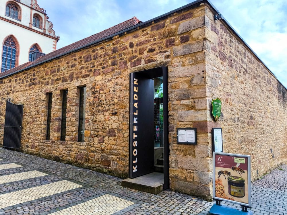 Ein Steinbau mit schmalen Fenstern und einem Schild mit der Aufschrift KLOSTERLADEN über dem Eingang in Fulda. Draußen gibt es eine Schautafel mit Produktbildern und einen Kopfsteinpflasterweg im Vordergrund.