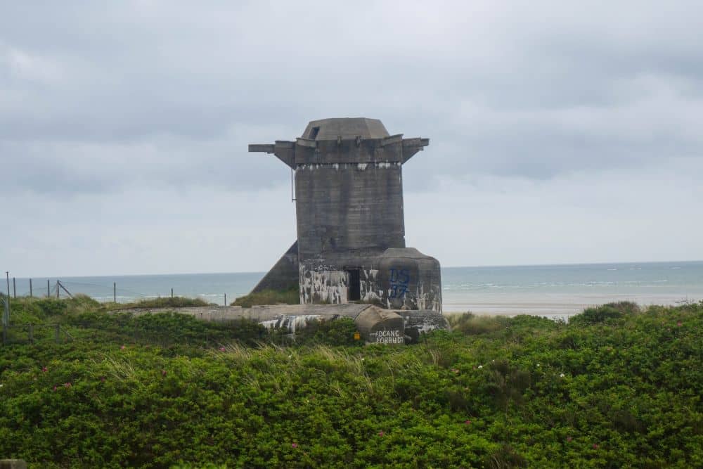 Ein Betonbunker aus dem Zweiten Weltkrieg steht wildromantisch auf einer grasbewachsenen Düne bei Blavand, Dänemarks Westen, mit dem Meer und dem bewölkten Himmel im Hintergrund. Das verwitterte Bauwerk ist mit Graffiti beschmiert.