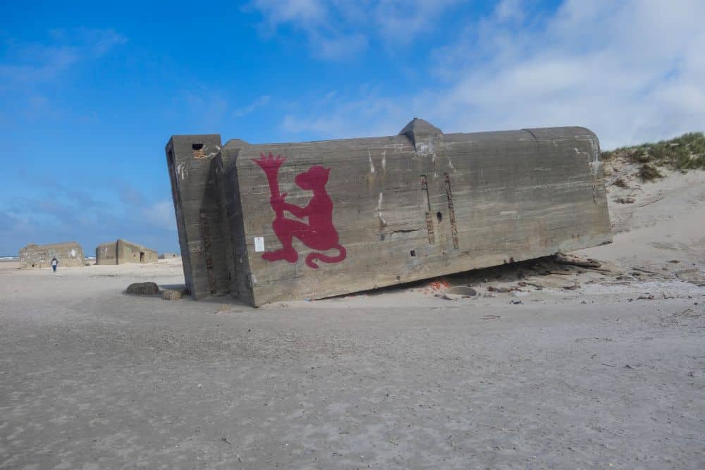 Ein großer Betonbunker liegt gekippt auf dem wildromantischen Sandstrand von Blavand in Dänemarks Westen, bedeckt mit einem Graffiti eines roten Löwen, der eine Krone hält. Grasbewachsene Dünen und ein blauer Himmel mit Wolken vervollständigen die Szene, während in der Ferne eine Person spazieren geht.