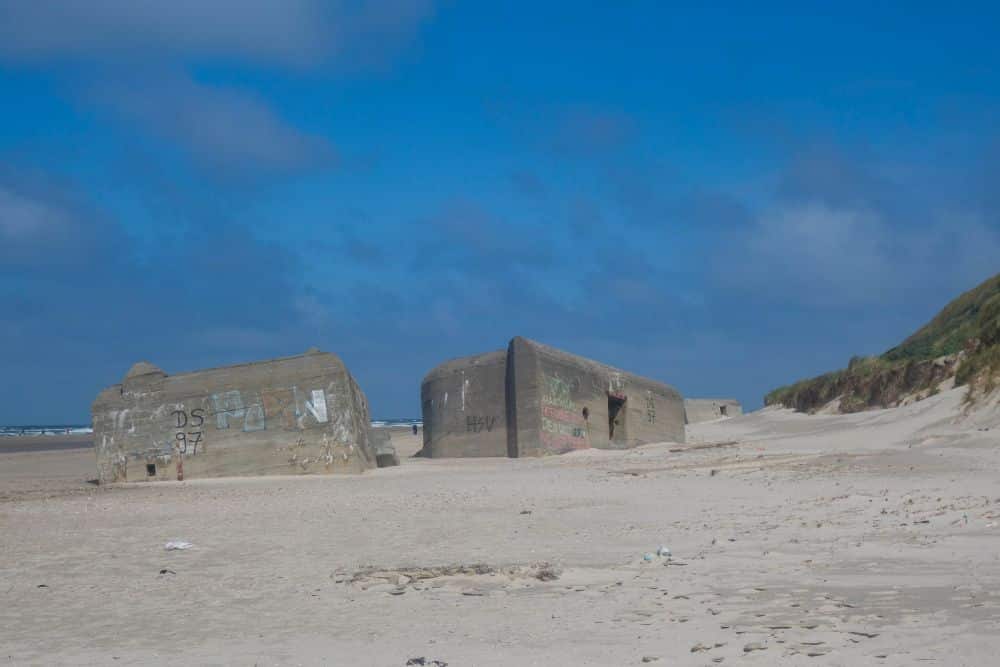 Große Betonbunker, teilweise im Sand vergraben, liegen an einem verlassenen Strand unter Dänemarks Westens wildromantisch blauem Himmel. An den Bunkern sind einige Graffiti zu sehen, und auf der rechten Seite bei Blavand erheben sich grasbewachsene Dünen.