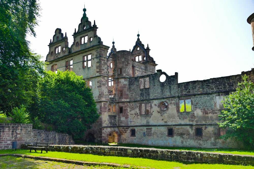 In Hirsau steht eine historische Steinburg mit verzierten Giebeln und hohen Türmen, die teilweise verfallen ist, umgeben von grünen Bäumen und Gras, wobei das Sonnenlicht Teile des Bauwerks beleuchtet.