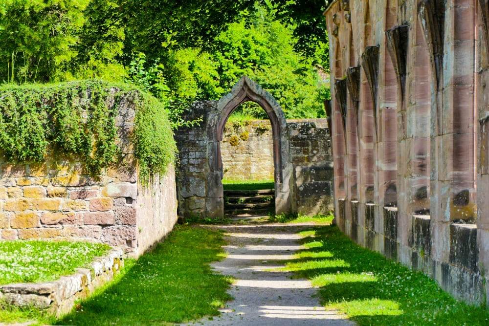 Ein sonnenbeschienener, von Gras gesäumter Steinweg führt durch ein bogenförmiges Steintor in den alten Steinmauern von Hirsau, umgeben von üppig grünen Bäumen im Hintergrund.