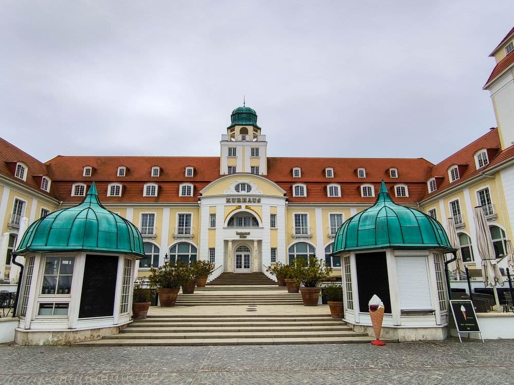Ein prachtvolles Hotel mit rotem Dach und grünen Kuppeln in Binz, mit einem bogenförmigen Eingang, der mit "Kurhaus" beschriftet ist. Eine breite Treppe führt zum Haupteingang, flankiert von grün überdachten Pavillons und Topfpflanzen. Draußen steht eine riesige Eiswaffel.
