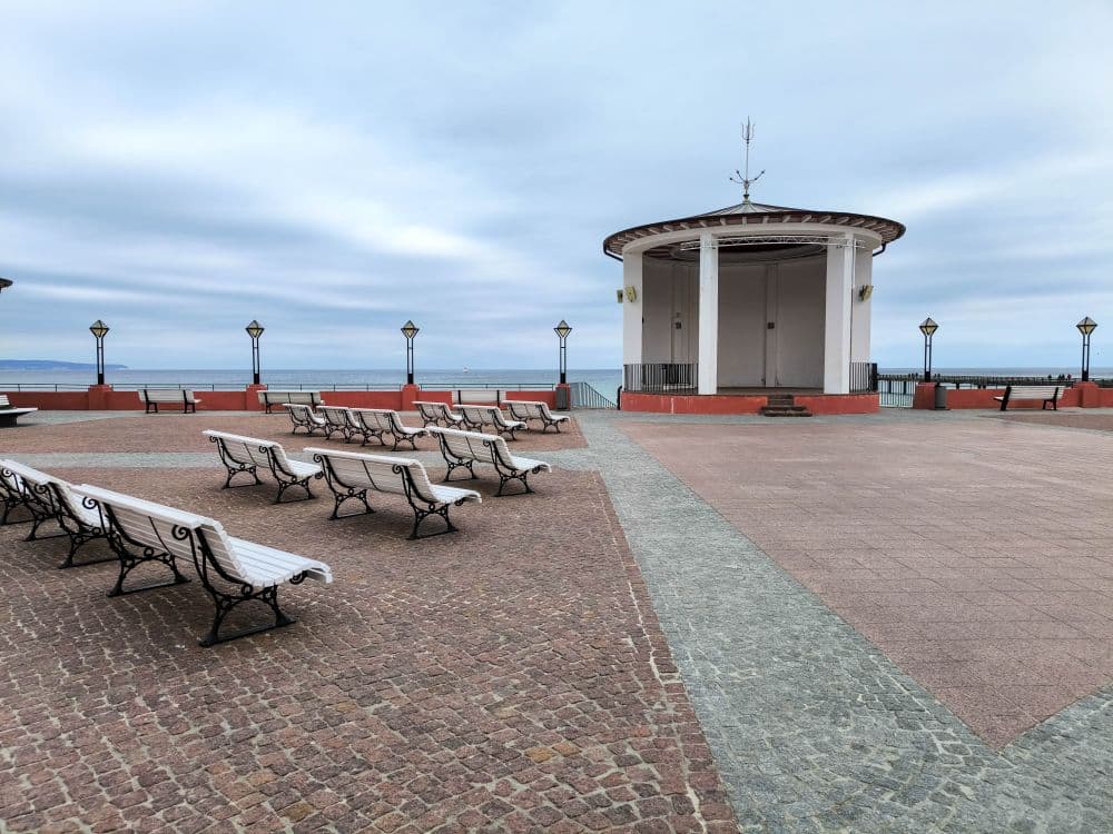 Freiluftpavillon am Meer in Binz mit weißen Bänken auf einem gepflasterten Platz; der Himmel ist bewölkt und das Meer ist im Hintergrund zu sehen.