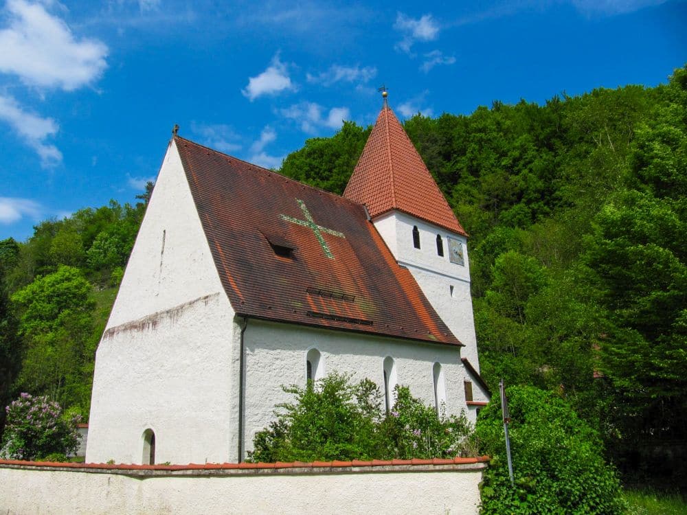 Eine kleine weiße Kirche mit einem steilen roten Ziegeldach und einem quadratischen Turm steht inmitten grüner Bäume im Kleinen Lautertal und bietet eine friedliche Auszeit unter einem strahlend blauen Himmel mit vereinzelten Wolken.