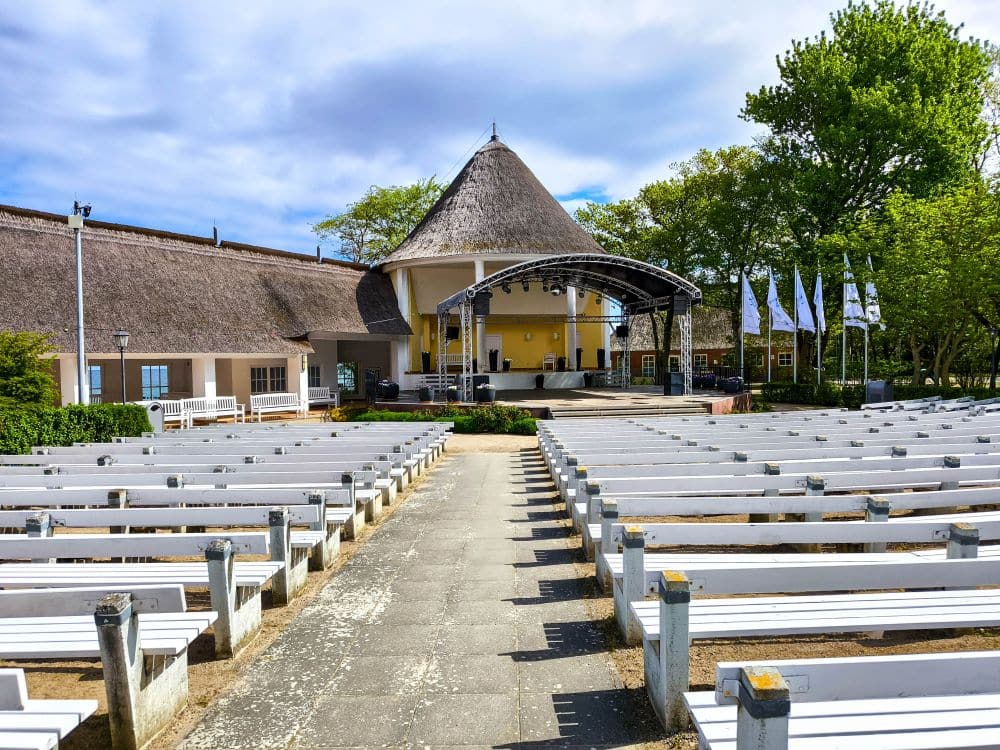 Freiluft-Amphitheater mit weißen Holzbänken vor einer Bühne unter einem gewölbten Vordach, umgeben von Bäumen und Fahnenmasten in Kühlungsborn. Genießen Sie eine Auszeit an einem sonnigen Tag, mit einem reetgedeckten Gebäude hinter der Bühne.