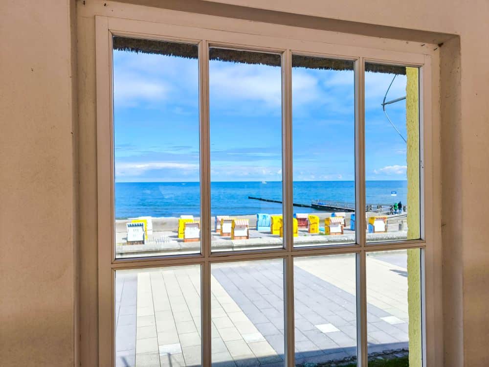 Blick durch ein Fenster auf eine sonnige Ostsee-Strandszene in Kühlungsborn, mit bunten Strandkörben, einem gepflasterten Weg und einer Seebrücke, die sich in das strahlend blaue Meer erstreckt - perfekt für eine Auszeit bei teilweise bewölktem Himmel.