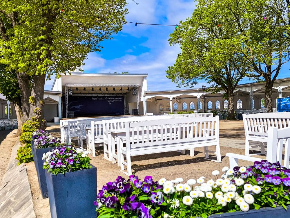Weiße Bänke und Blumenkübel mit lila und weißen Blumen stehen vor einer Freilichtbühne in Kühlungsborn und bieten eine entspannende Auszeit inmitten großer grüner Bäume unter dem blauen Ostseehimmel.