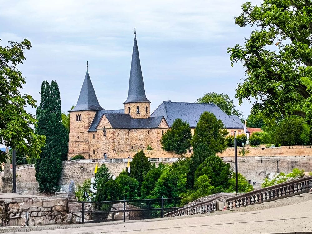 Eine historische Steinkirche mit zwei hohen, spitzen Türmen steht inmitten üppiger Bäume und Grünflächen unter einem bewölkten Himmel in Fulda. Im Vordergrund sind eine Steinmauer und ein abschüssiger Weg zu sehen.