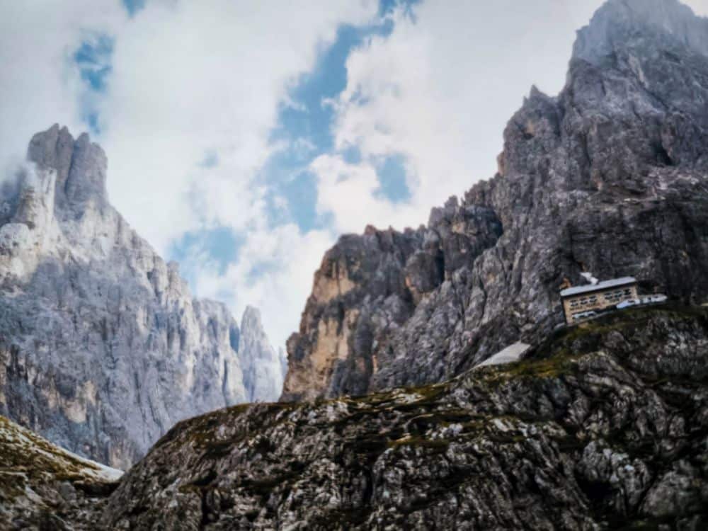 Eine zerklüftete Berglandschaft mit dramatischen Felsgipfeln unter einem teilweise bewölkten Himmel; ein kleines Gebäude oder eine Hütte thront am Hang rechts und fängt die majestätische Schönheit der Dolomitenklassiker bei der Langkofelscharte ein.