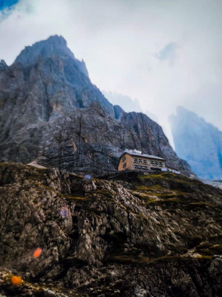 Ein steinernes Gebäude mit mehreren Fenstern steht auf einem felsigen Berghang in den Dolomiten, umgeben von steilen, zerklüfteten Felsen und nebligen Wolken in einer dramatischen, rauen Landschaft.