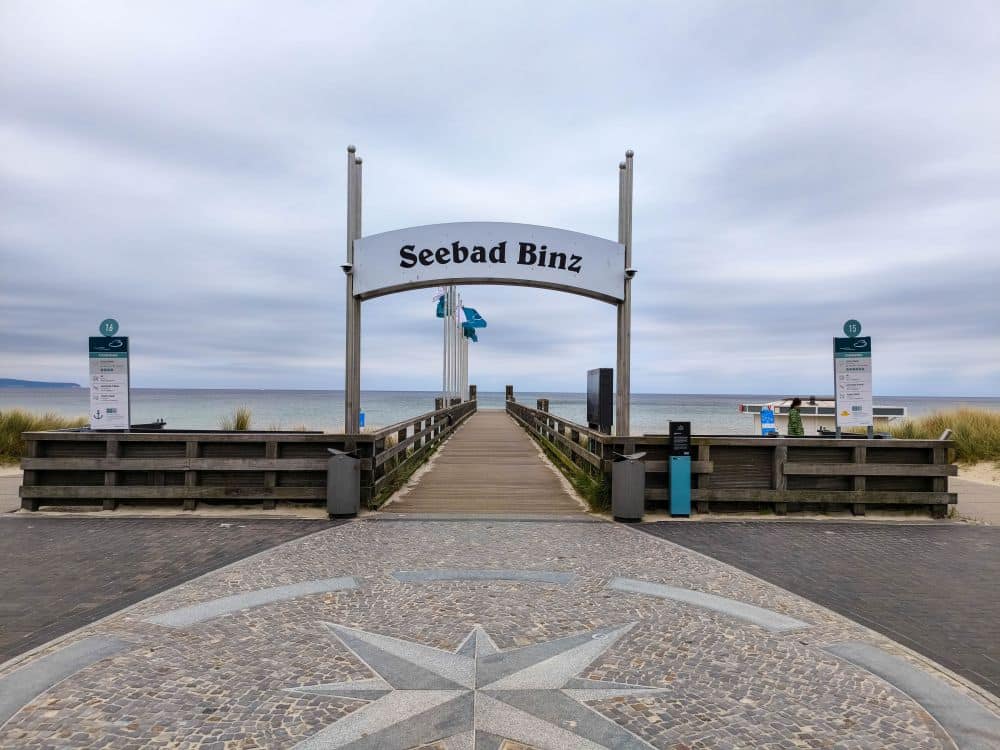 Unter einem wolkenverhangenen Himmel erstreckt sich eine hölzerne Seebrücke mit der Aufschrift Binz in Richtung Meer, mit Schildern, Geländern und Strandgras auf beiden Seiten und einer Mosaik-Kompassrose auf dem Gehweg davor.