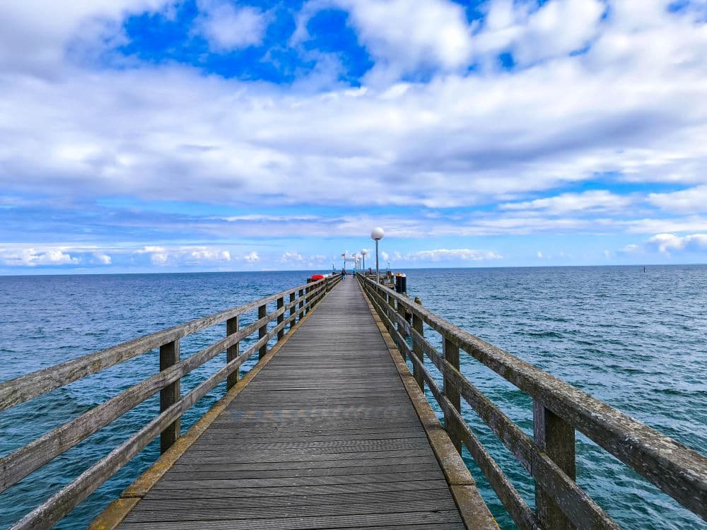 Eine hölzerne Seebrücke in Kühlungsborn ragt bei leicht bewölktem Himmel in die ruhige, blaue Ostsee hinaus, mit Geländern auf beiden Seiten und sichtbaren Lampen am Ende - perfekt für eine entspannte Auszeit am Meer.