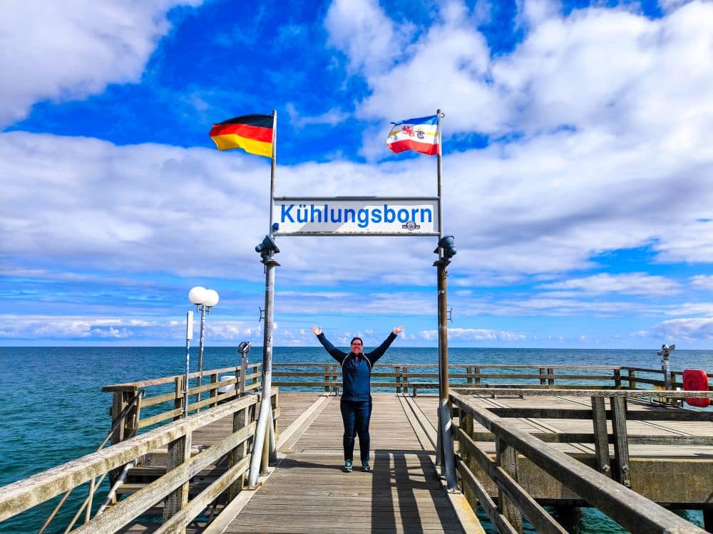 Bei teilweise bewölktem Himmel steht eine Person mit erhobenen Armen auf einem Holzsteg über der Ostsee. Darüber steht ein Schild mit der Aufschrift Kühlungsborn, und es wehen zwei Flaggen, eine deutsche und eine regionale - ein perfekter Ort für eine Auszeit.