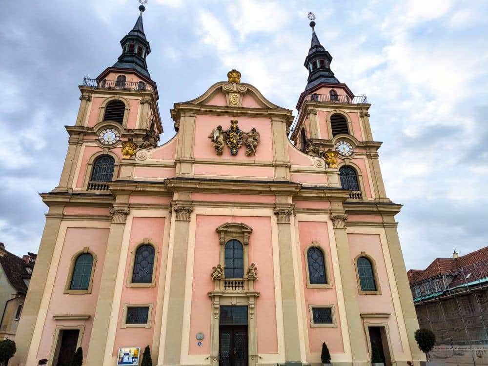In Ludwigsburg erhebt sich eine prächtige Barockkirche mit zwei hohen Glockentürmen, kunstvollen Details, Bogenfenstern und einer blassrosa-gelben Fassade, in der sich die Barocke Pracht vor einem wolkenverhangenen Himmel spiegelt.