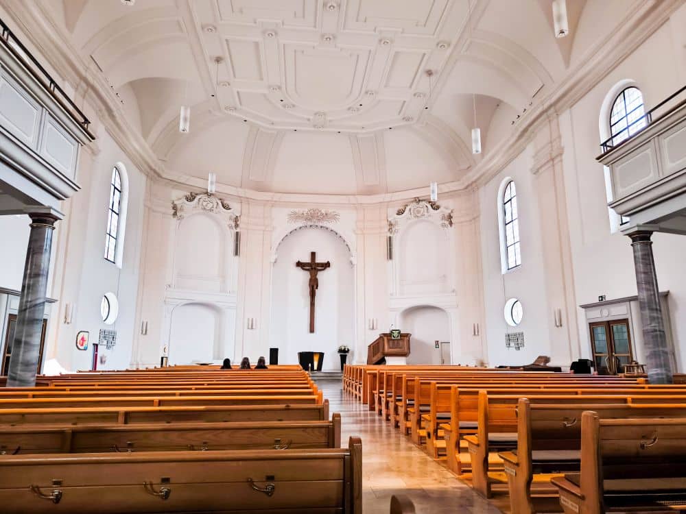 Innenraum einer hellen Barockkirche in Ludwigsburg mit hölzernen Kirchenbänken, einer verzierten hohen Decke, Bogenfenstern und einem großen Kruzifix über dem Altar. Ein paar Leute sitzen in der Nähe der Vorderseite und genießen die heitere Atmosphäre.
