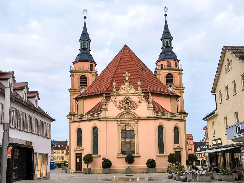 Barocke Pracht: Barocke Kirche mit zwei Türmen und rotem Ziegeldach auf einem Ludwigsburger Stadtplatz, umgeben von Gebäuden, Geschäften, Bunten Gärten in Pflanzkübeln und kleinen Bäumen. Der Himmel ist bewölkt.