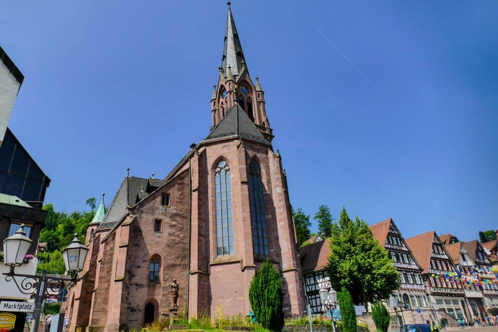Eine hohe, historische Kirche mit spitzem Kirchturm und großen Bogenfenstern steht neben traditionellen Fachwerkhäusern in Calw unter einem klaren blauen Himmel. Das helle Sonnenlicht hebt die Steinfassade des Gebäudes und die umliegenden Grünflächen hervor.