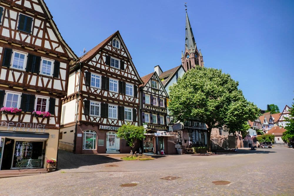 Ein sonniger Stadtplatz in Calw mit traditionellen Fachwerkhäusern, einer Kirche mit einem hohen Turm, Bäumen, Ladenschildern und Kopfsteinpflaster in einem malerischen europäischen Dorf.