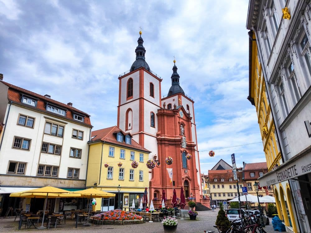 Ein lebendiger Stadtplatz in Fulda mit bunten Gebäuden, Straßencafés und einer großen rot-weißen Kirche mit Zwillingstürmen unter einem teilweise bewölkten Himmel. Hängende Dekorationen und Blumen verleihen der lebendigen Szene Charme.