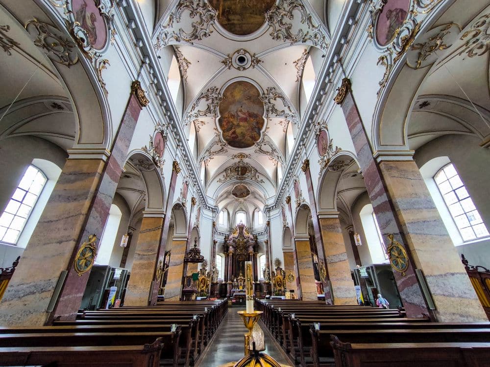 Innenraum einer großen Kirche in Fulda mit kunstvollen Gewölbedecken, dekorativen Bögen, detaillierten Fresken und Reihen von Holzbänken, die zu einem kunstvollen Altar führen. Durch die hohen Bogenfenster fällt Licht ein, das die architektonische Pracht hervorhebt.