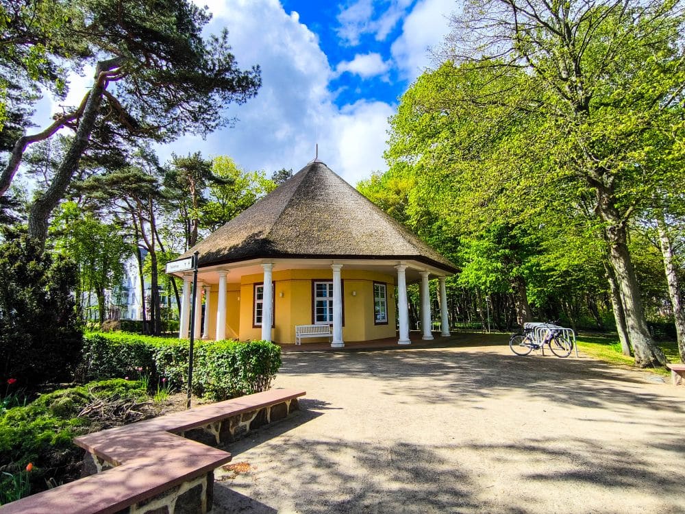 Ein kleines gelbes Reetdachhaus mit weißen Säulen steht in einem sonnigen, baumbestandenen Park in Kühlungsborn an der Ostsee. In der Nähe sind Fahrräder abgestellt, und Bänke säumen einen Sandweg unter einem teilweise bewölkten blauen Himmel - perfekt für eine Auszeit.