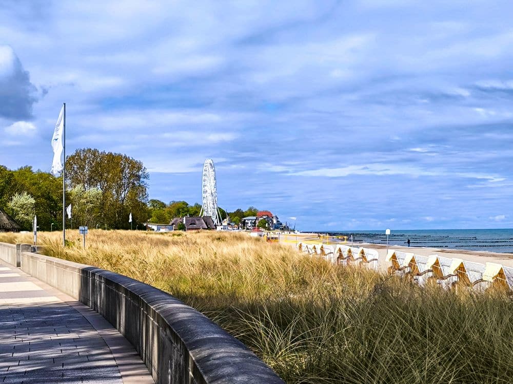 Eine von hohem Gras gesäumte Uferpromenade in Kühlungsborn führt zu einem mit Strandkörben gesäumten Sandstrand der Ostsee. In der Ferne sind ein Riesenrad und Gebäude zu sehen, und der Himmel ist teilweise bewölkt - perfekt für eine Auszeit.