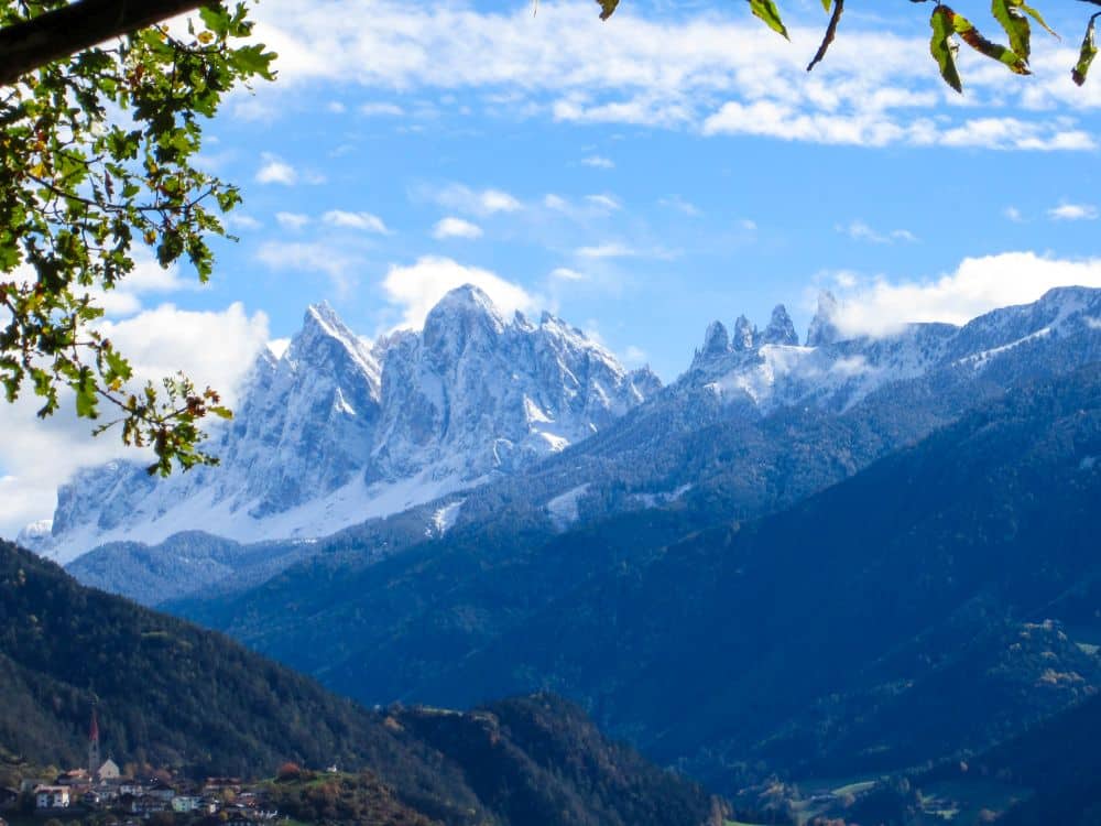 Schneebedeckte Berggipfel erheben sich über grünen Hügeln unter einem blauen Himmel mit vereinzelten Wolken; Baumzweige umrahmen den oberen Bildrand und fangen die landschaftliche Schönheit entlang des Keschtnwegs bei Feldthurns in Südtirol ein.