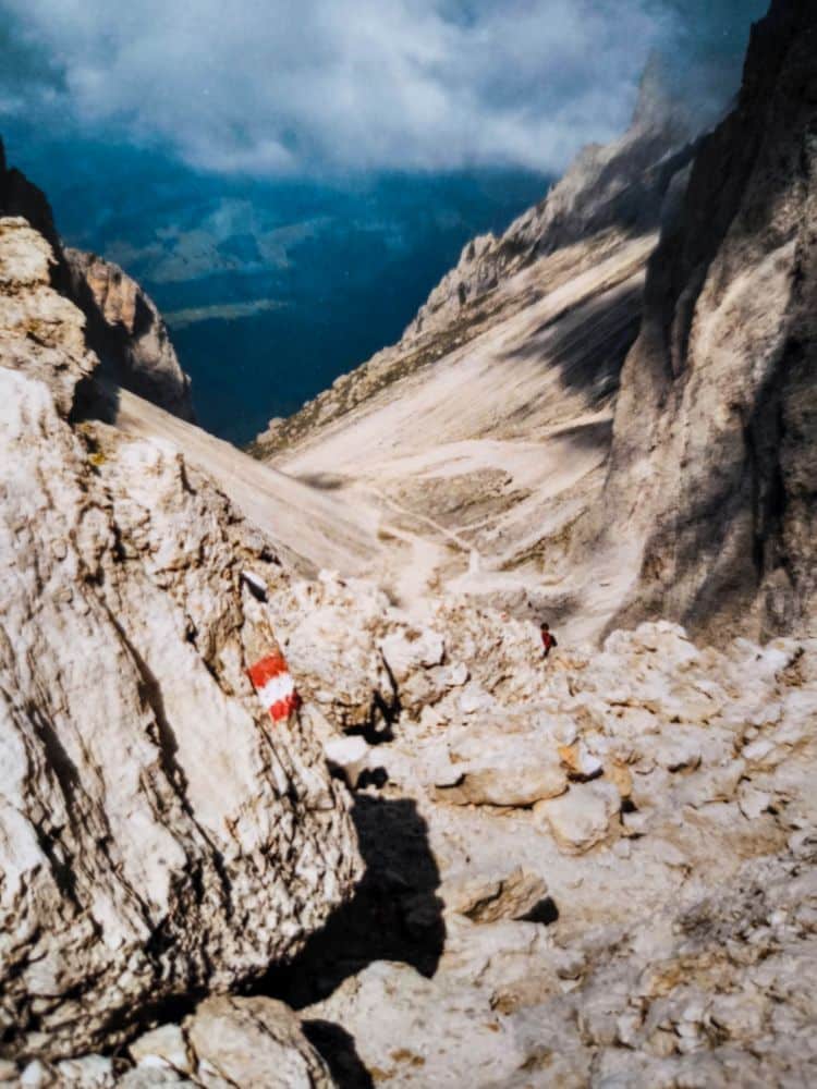 Ein felsiger Bergpass in den Dolomiten mit schroffen Felsen und einem schmalen Pfad, der sich durch die Langkofelscharte schlängelt. Auf einem Felsen im Vordergrund ist bei bewölktem Himmel eine rot-weiße Wegmarkierung zu sehen.