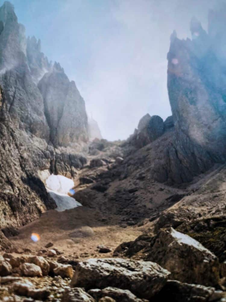 Felsige Berglandschaft mit zerklüfteten Gipfeln, einem zerklüfteten Pfad und Schneeflecken unter einem dunstigen Himmel. Das raue Terrain erinnert an die dramatischen Dolomitenklassiker bei der Langkofelscharte in den legendären Dolomiten.