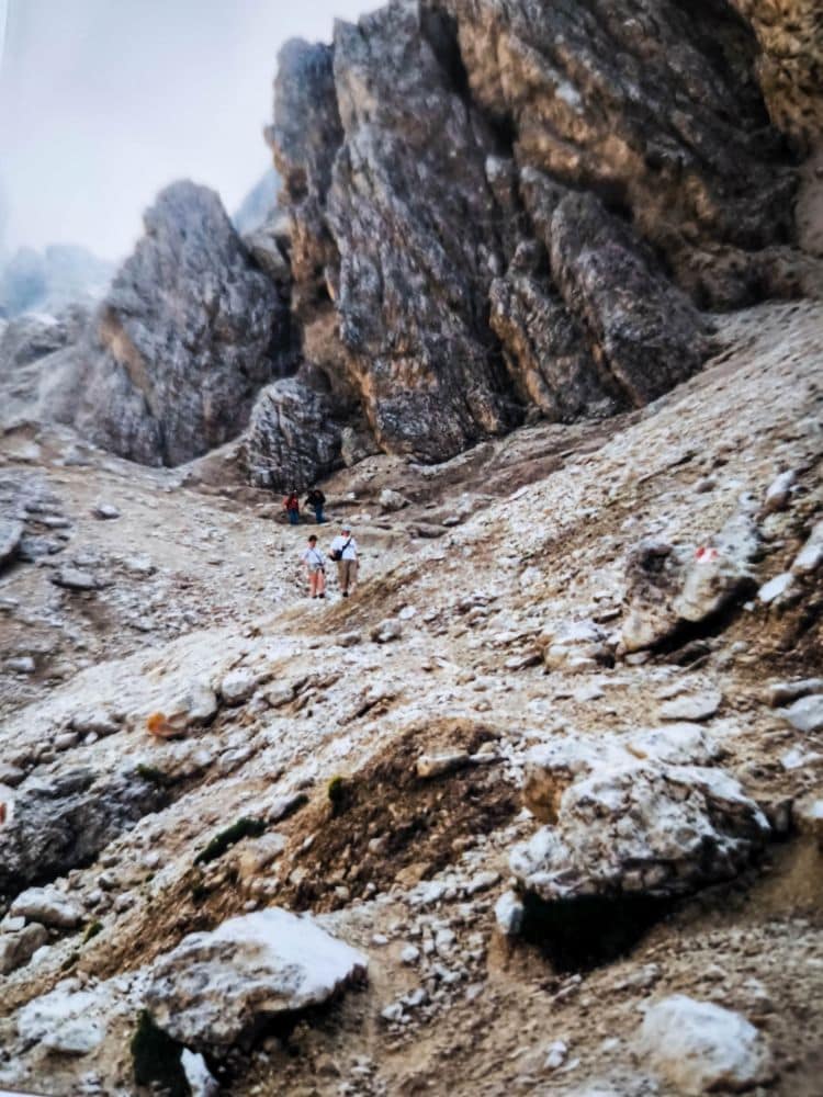 Drei Wanderer steigen bei bewölktem Himmel einen schroffen Dolomitenhang mit steilen, zerklüfteten Felsen und spärlicher Vegetation hinauf. Im Hintergrund ragt die dramatische Langkofelscharte auf, die die wilde Schönheit dieser Dolomitenklassiker-Landschaft noch verstärkt.
