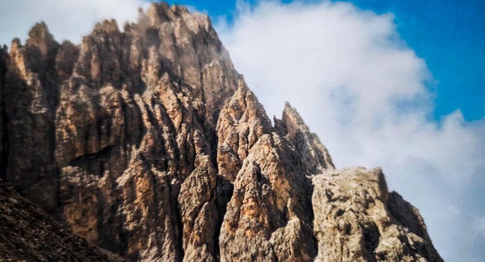 Die zerklüfteten Felsgipfel der Dolomitenklassiker ragen steil in den strahlend blauen Himmel, und weiße Wolkenfetzen verdecken teilweise die Langkofelscharte. Die schroffe Struktur der Felsen wird durch das Sonnenlicht wunderschön hervorgehoben.