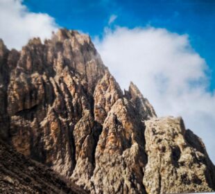 Die zerklüfteten Felsgipfel der Dolomitenklassiker ragen steil in den strahlend blauen Himmel, und weiße Wolkenfetzen verdecken teilweise die Langkofelscharte. Die schroffe Struktur der Felsen wird durch das Sonnenlicht wunderschön hervorgehoben.